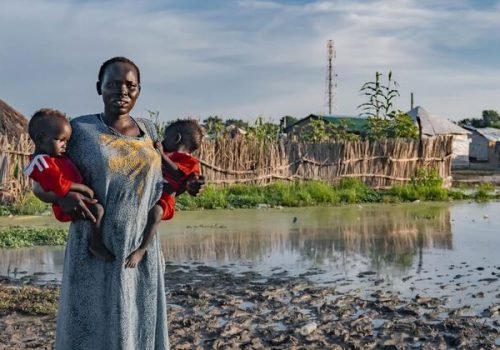© Unicef
                          Uma mulher segura seus filhos em frente à sua casa atingida pela enchente no Sudão do Sul